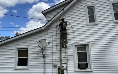 A person on a ladder repairing exterior cables on a white house under a blue sky.