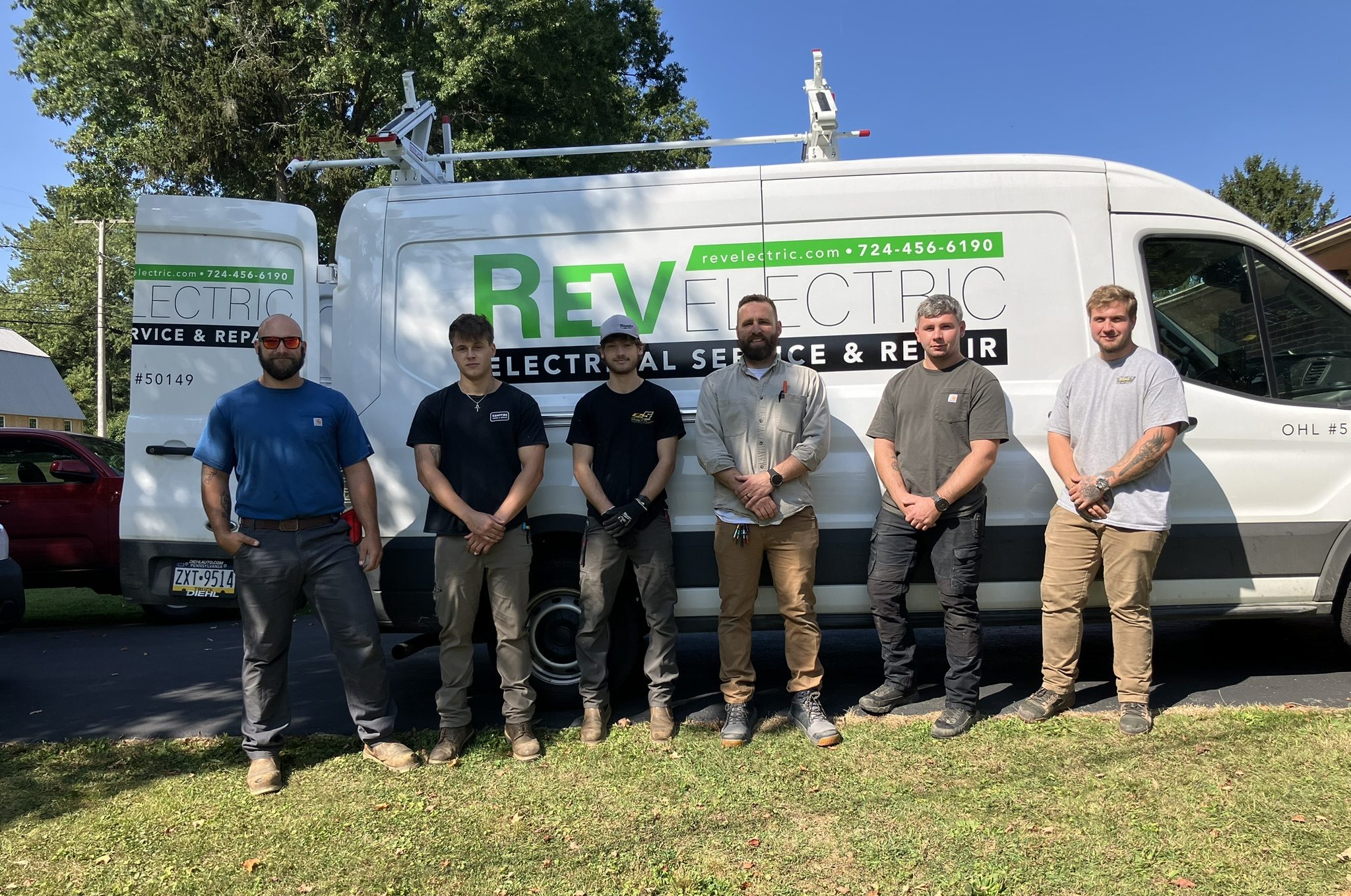 Six electricians standing in front of a white Rev Electric service van displaying logo and phone number