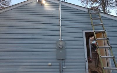 Blue vinyl-sided house with electrical meter and service mast conduit, ladder at doorway and a person working on the roof.