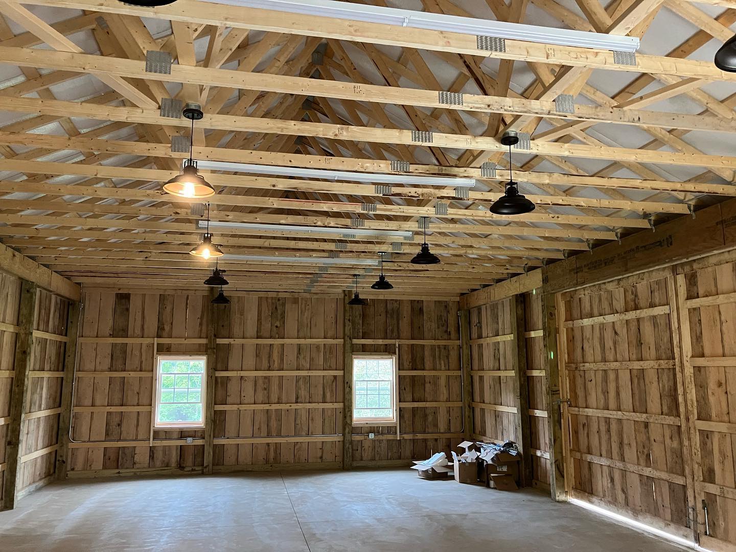 Interior of wooden barn/workshop with exposed roof trusses, hanging pendant lights, concrete floor and two windows