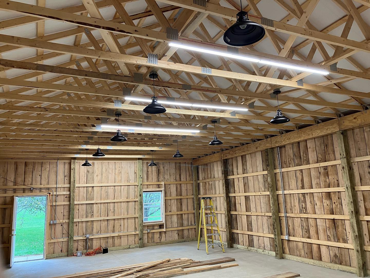 Unfinished wooden workshop interior with exposed trusses, pendant lights, ladder, stacked lumber and concrete floor