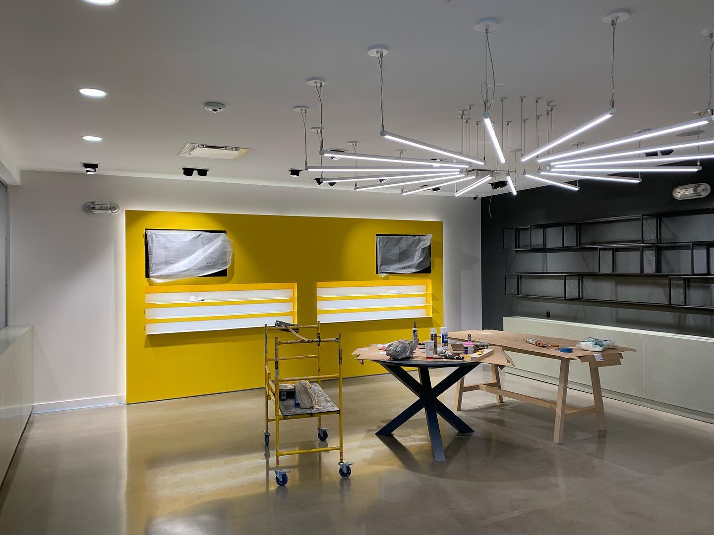 Modern retail space under construction with yellow display wall, suspended linear lights, tables, scaffolding, and shelving.