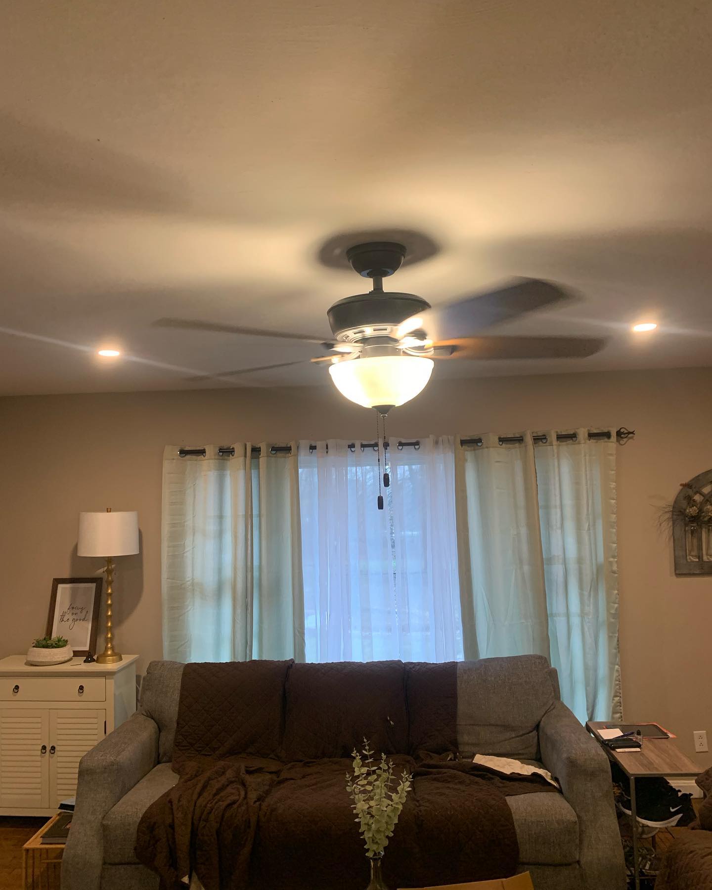 Cozy living room interior with lit ceiling fan, gray sofa, sheer curtains, side lamp and decorative accents