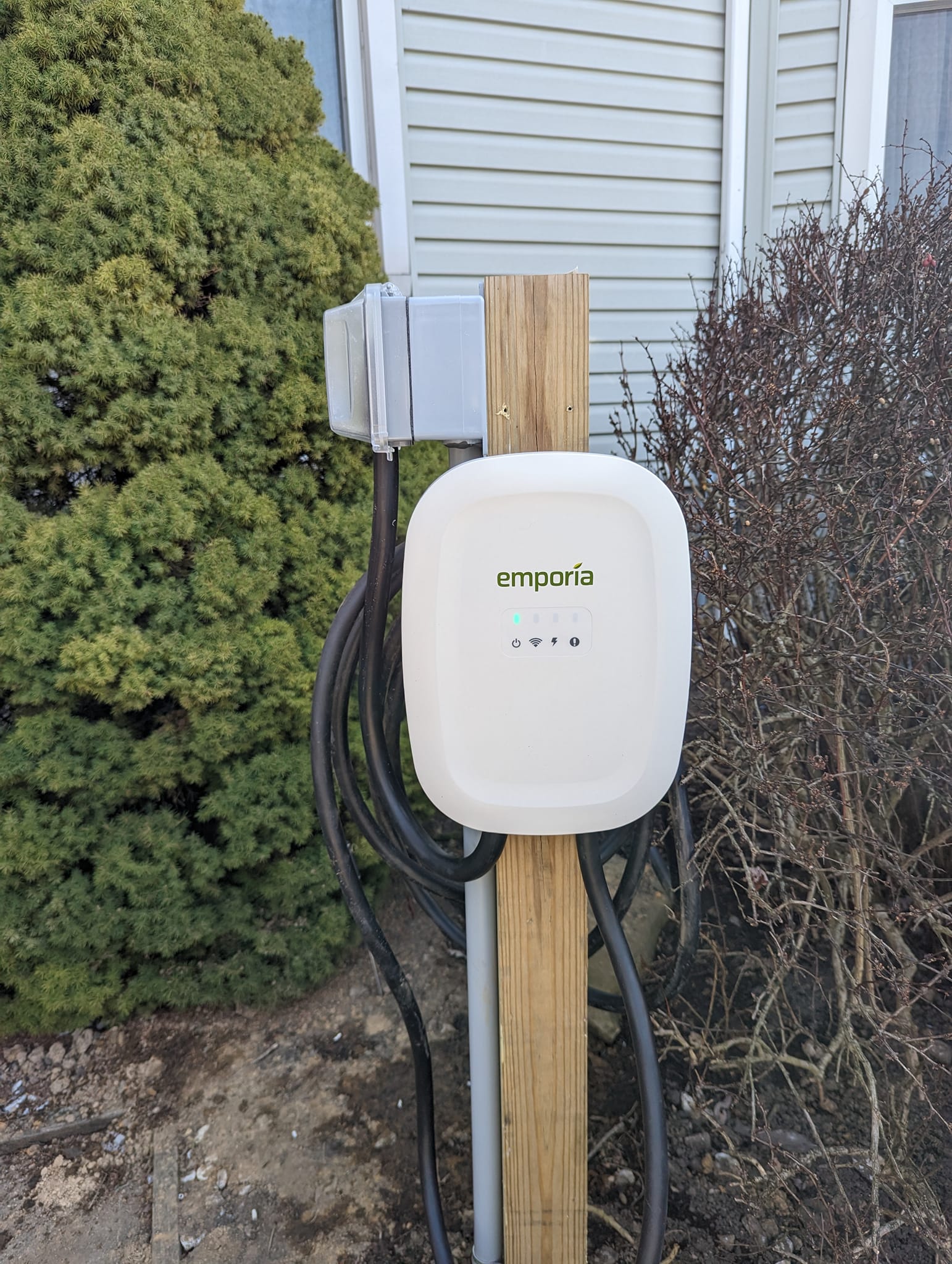 Emporia electric vehicle charger mounted on a wooden post with coiled cables next to house siding and shrubs