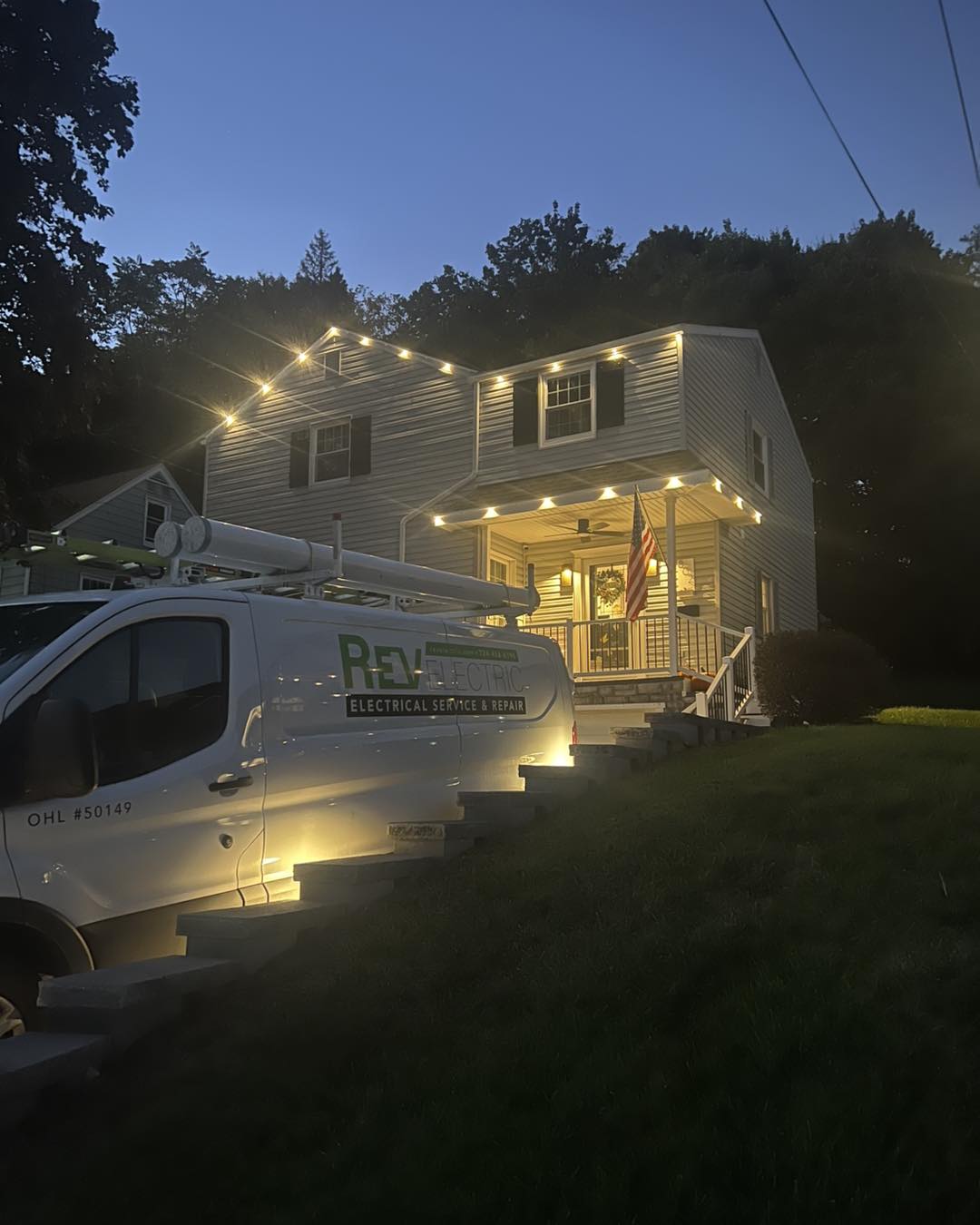 Two-story house outlined by warm string lights, illuminated porch and Rev Electric service van in driveway.