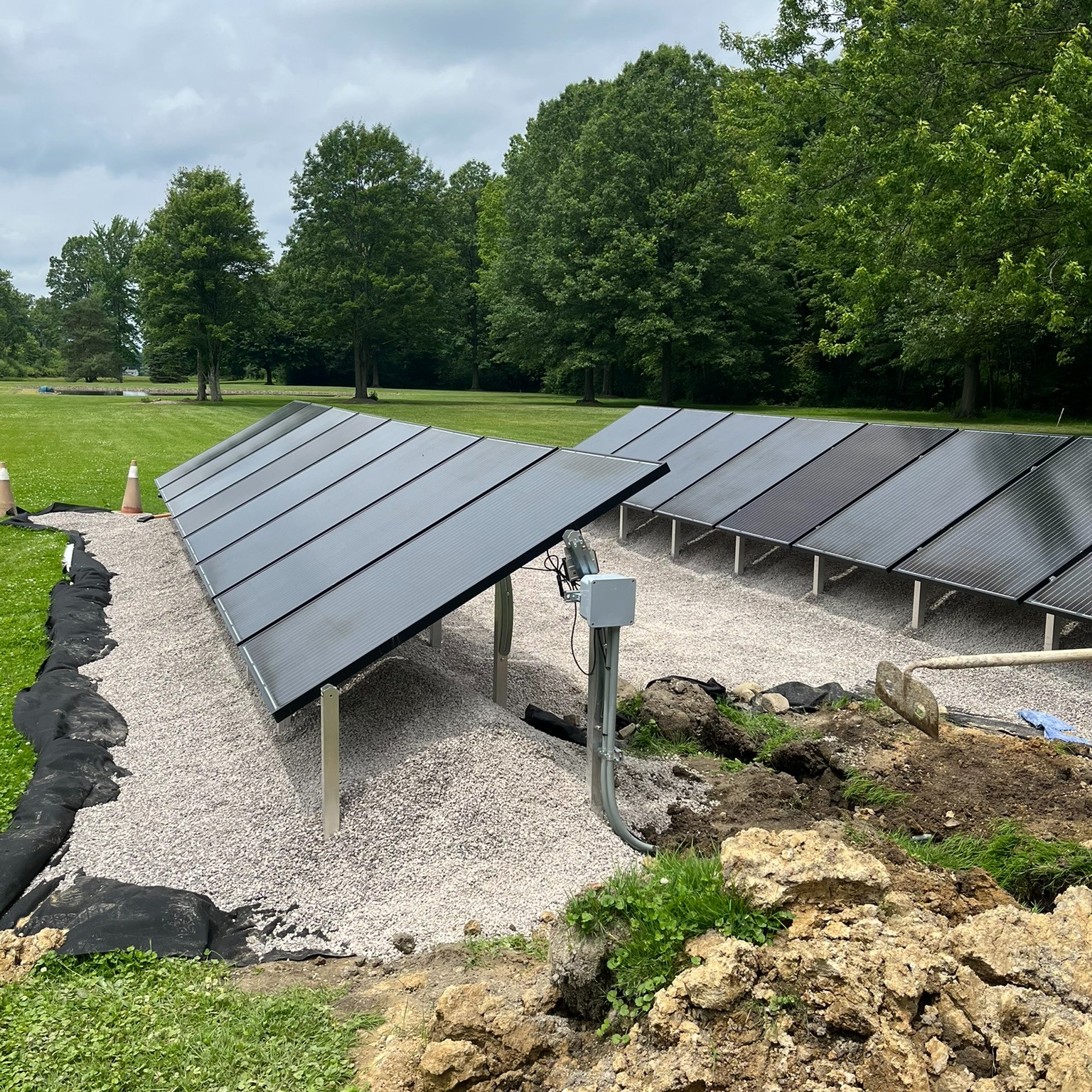 Ground-mounted solar panels on gravel in a grassy field with trees and an overcast sky
