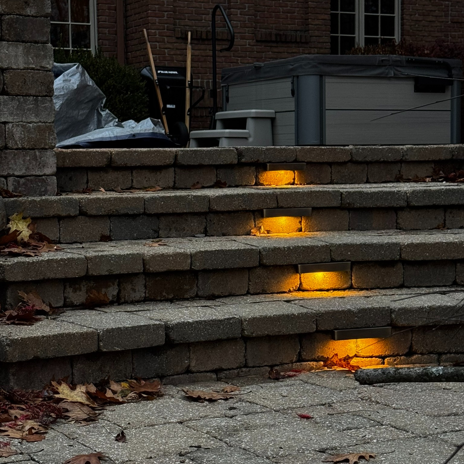 Outdoor stone steps with recessed amber LED lights, fallen leaves and a hot tub in the background at dusk.