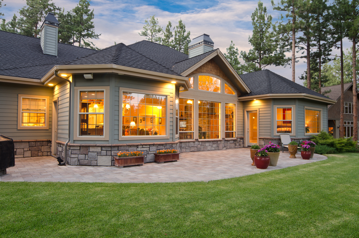 Modern home exterior at dusk with large lit windows, stone-trim patio, potted flowers and manicured lawn