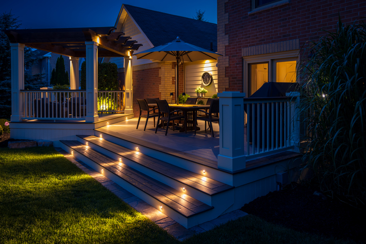 Cozy illuminated backyard deck at dusk with built-in step lights, dining table, umbrella, and pergola by brick house