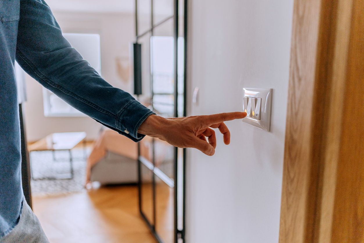 A person in a denim shirt reaching to press a wall light switch in a modern living room doorway