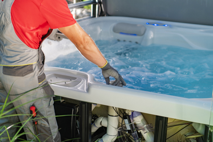 Technician in gloves servicing a bubbling hot tub, inspecting exposed plumbing and equipment under the spa panel