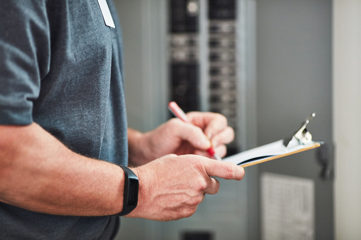 Person writing on a clipboard while inspecting an electrical panel, wearing a wrist fitness tracker.